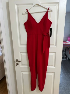 Röd jumpsuit  - Snygg röd jumpsuit från Trixxi med smala axelband, v-ringning och ett matchande tygskärp i midjan. Jumpsuiten har en figurnära överdel och raka ben🫶🏼