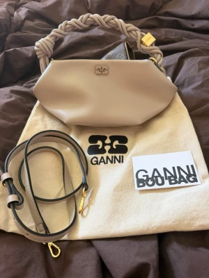 Ganni bou bag❤️ - Snygg beige handväska från Ganni med flätat handtag och avtagbar axelrem i läder. Väskan har guldfärgade detaljer och stängs med magnetknapp. Perfekt accessoar för dig som vill ha en trendig och stilren look.