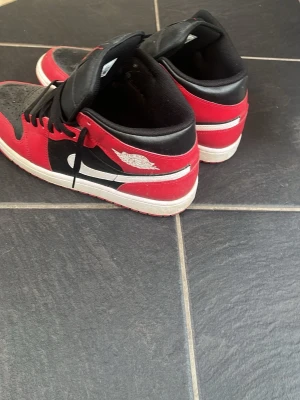 Svarta och röda Air Jordan 1 Mid - Säljer ett par Air Jordan 1 Mid sneakers i svart, rött och vitt. Skorna har klassisk Nike Swoosh på sidan, svarta skosnören och perforerad tåbox. Materialet är en mix av läder och syntet. Ikonisk Air Jordan-logga på sidan och Jumpman på plösen.