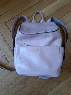 Ljusrosa ryggsäck i konstläder - Snygg ljusrosa ryggsäck i konstläder med enkel design och silverfärgade dragkedjor. Ryggsäcken har ett stort huvudfack, en framficka med dragkedja och justerbara axelband. Perfekt för dig som vill ha en stilren och praktisk väska till vardagen.