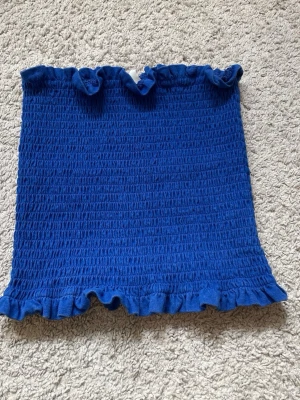 Blå bandeau tubtopp med volang - Säljer en blå tubtopp med smockad struktur och volangdetaljer upptill och nedtill. Toppen är ärmlös och har en tight passform som sitter snyggt på kroppen. Perfekt för sommardagar eller festivaler!