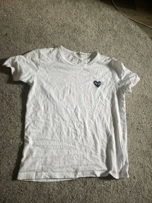 Vit t-shirt Comme des Garçons Play - Vit t-shirt från Comme des Garçons Play med det ikoniska svarta hjärta-trycket på bröstet. Klassisk rund hals och korta ärmar. Tillverkad i mjuk bomull som känns skön mot huden. Perfekt för en clean och stilren look.