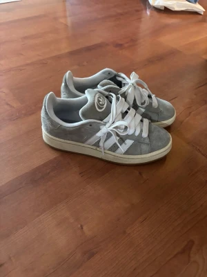 Grå Adidas Campus sneakers - Säljer ett par grå Adidas Campus sneakers i mocka med vita detaljer och klassiska tre ränder på sidan. Skorna har vita skosnören och en beige sula. Perfekta för dig som gillar en clean och stilren look. 