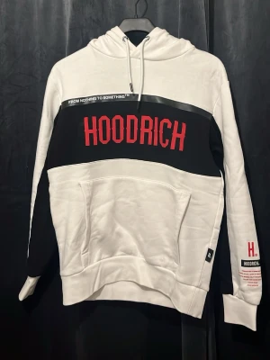 Vit och svart hoodie från Hoodrich - Snygg hoodie i vitt och svart från Hoodrich med stor röd logga på bröstet och textdetaljer. Hoodien har huva med dragsko, stor magficka och kontrastpaneler på ärmarna. Materialet är mjukt och skönt, perfekt för en chill och street-inspirerad look.