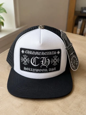 Chrome Hearts svartvit truckerkeps - Säljer en svartvit truckerkeps från Chrome Hearts med broderad patch framtill där det står 'Chrome Hearts Hollywood, USA' och CH-logga. Kepsen har mesh på sidorna och bak, samt en silverfärgad detalj på toppen. Justerbar baktill och klassisk rak skärm.