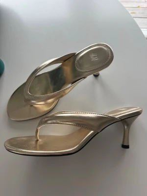 Guldiga klackade sandaler från H&M - Snygga guldiga sandaler från H&M med smal klack och tårem. De har en metallic finish och öppen design som ger en trendig look. Perfekta för dig som vill sticka ut med ett par glammiga skor. Materialet ser ut att vara konstläder.