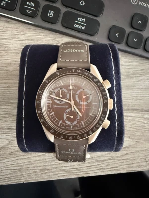 Omega x Swatch Speedmaster brun klocka - Snygg brun Omega x Swatch Speedmaster med tachymeter och kronograf. Klockan har ett brunt armband i textil med vita sömmar och logga, samt en rund urtavla med flera små urtavlor och tydliga markeringar. Perfekt för dig som gillar stilrena accessoarer.