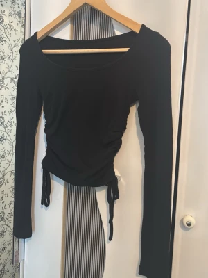 Svart långärmad topp med snörning - Snygg svart långärmad topp med rund halsringning och trendig snörning i sidorna som ger en cool, croppad look. Tillverkad i mjukt material som sitter skönt på kroppen. Perfekt att styla med jeans eller kjol för en modern vibe.