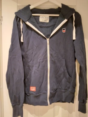 Mörkblå hoodie med dragkedja, strlk Small - Mörkblå hoodie med vit dragkedja och vita snören i huvan. Två stora fickor framtill och små broderade detaljer på bröstet och fickan. Mjuk bomullskänsla och ribbade muddar vid ärmar och nederkant. Stor röd detalj på huvan baktill. Strlk Small. Bra skick - se bilder för egen bedömning.