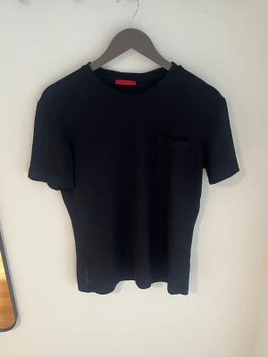 Svart Prada t-shirt med bröstficka - En stilren svart Prada t-shirt i bomull med rund hals och en liten bröstficka på vänster sida. Klassisk slim fit passform och korta ärmar.