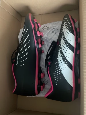 Adidas Predator svart/vita fotbollsskor med ben skydd - Säljer ett par Adidas Predator fotbollsskor i svart med vita och rosa detaljer. Skorna har klassisk snörning, dobbar för gräsplan och en modern design med mönstrade sidor. Perfekta för dig som vill ha stilrena och funktionella skor på planen.