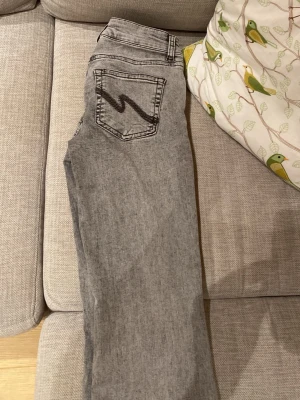 Grå raka jeans med bakficka - Säljer ett par grå boucut jeans med rak passform och klassisk femficksdesign. Jeansen har en tydlig dekorsöm på bakfickan i form av ett Z-mönster. Materialet är denim i en ljusgrå tvätt som passar till det mesta. Perfekta för en avslappnad och trendig look.