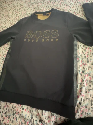 Svart sweatshirt från Hugo Boss - Snygg svart sweatshirt från Hugo Boss med stort BOSS-tryck i guld på bröstet. Tröjan har rund halsringning och ribbade muddar. Perfekt för en clean och stilren look. Materialet känns mjukt och bekvämt mot huden.