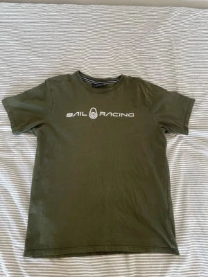 Olivgrön Sail Racing t-shirt - Snygg olivgrön t-shirt från Sail Racing med vit logga tryckt på bröstet och en mindre logga i nacken. Klassisk passform och rund halsringning. Perfekt till jeans eller shorts för en avslappnad stil.