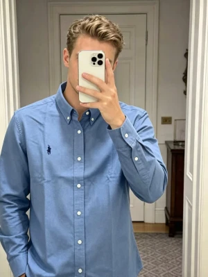 Blå skjorta från Ralph Lauren - Klassisk blå skjorta från Ralph Lauren med button-down krage och vita knappar. Skjortan har lång ärm och den ikoniska loggan broderad på bröstet. Perfekt till jeans eller chinos för en clean och stilren look.
