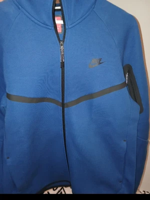 Nike Tech Fleece hoodie blå & svart - Två Nike Tech Fleece hoodies i blått och svart med hel dragkedja, huva och fickor med dragkedja på ärmen. Snygg design med diskret Nike-logga på bröstet och en vågig detalj över bröstet. Perfekt för en sportig och avslappnad stil. Blåa helt ny köpt fel o tappat kvittot köptes för 1399 st den svarta är sånt gott som.ny med använd 4 ggr max  900 för blåa  st M och svarta 800 st m