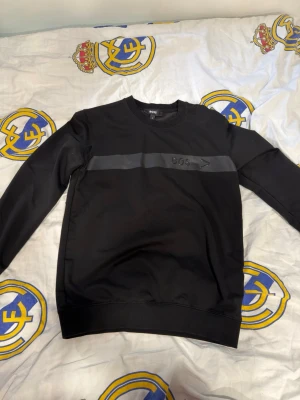 Svart sweatshirt från BOSS - Svart sweatshirt från BOSS med rund halsringning och en diskret mörkgrå rand över bröstet där loggan är placerad. Tröjan har långa ärmar och ribbade muddar vid ärmslut och nederkant. Perfekt för en stilren och enkel look. Tyvärr har ena S’t fallit av i tvätten, där av det låga priset. Annars helt fläckfri.
