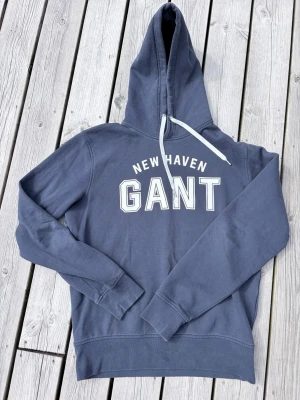 Blå hoodie från GANT med tryck - Säljer en blå hoodie från GANT med vit text 'NEW HAVEN GANT' på bröstet. Tröjan har huva med dragsko och mjukt, borstat foder på insidan. Klassisk passform och ribbade muddar vid ärmar och nederkant. Perfekt för en avslappnad stil.