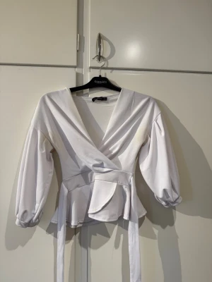Vit omlottblus från Boohoo - Säljer en snygg vit omlottblus från Boohoo med långa puffärmar och knytband i midjan. Blusen har djup v-ringning och en lätt figursydd passform som ger en trendig siluett. Perfekt att styla med jeans eller kjol för en chic look.