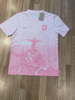 Vit och rosa t-shirt med Rio-motiv - Snygg t-shirt i vitt och ljusrosa med motiv av Kristusstatyn i Rio de Janeiro. T-shirten har korta ärmar, rund halsringning och Nike-logga på bröstet. Perfekt för dig som gillar unika tryck och streetstyle.