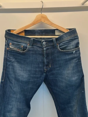 Mörkblå raka jeans med slitningar från Diesel  - Säljer ett par mörkblå jeans med raka ben och snygga slitningar. Jeansen har klassisk femficksdesign, kontrastsömmar och detaljer i kopparfärg. Materialet är kraftig denim i bomull. Perfekta för en avslappnad och trendig look.