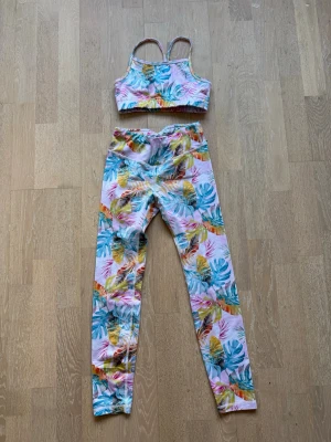Träningsset med tropiskt mönster - Säljer ett färgglatt träningsset med sport-bh och tights i tropiskt bladmönster. Setet har rosa bas med inslag av grönt, blått, orange och gult. 