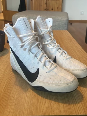 Vita höga sneakers från Nike - Säljer ett par vita höga sneakers från Nike med svart swoosh på sidan. Skorna har snörning hela vägen upp, ventilerande meshmaterial och förstärkta partier vid tån och hälen. Sulan är svart och ger bra grepp. Perfekta för dig som vill sticka ut med sportig stil.
