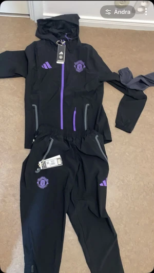 Manchester United träningsset svart/lila - Snyggt träningsset från Adidas med Manchester United-logga. Setet består av en svart jacka med huva och dragkedja samt matchande byxor. Lila detaljer och loggor ger en sportig look. Perfekt för träning eller chill. Materialet är lätt och andas.hjälper en vån att säja forbolls dressen 