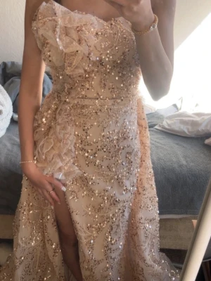 Champagnefärgad glittrig långklänning - Elegant långklänning i champagnefärg med glittriga paljetter och volangdetaljer. Klänningen har en axelbandslös design och en hög slits framtill. Perfekt för bal eller festliga tillfällen. Använts en gång till balen!