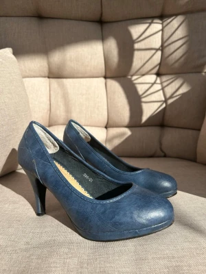 Pumps strl. 38 - Mörkblå pumps med rund tå och klassisk klack. Skorna har en stilren design i konstläder med lätt glansig finish och beige innersula med dekorativ kant. Lite sliten längst sömkanterna som syns på de två sista bilderna. Syns ej på långt håll.