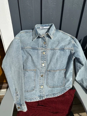 Ljusblå jeansjacka från Zara - Snygg ljusblå jeansjacka från Zara med klassisk krage, raka ärmar och stora framfickor. Jackan har silverfärgade knappar framtill och kontrastsömmar som ger en cool vintagekänsla. Perfekt att slänga över en hoodie eller t-shirt.
