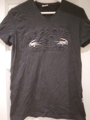 Svart Lacoste t-shirt med logo framtill, strlk Small - Svart t-shirt från Lacoste med broderad text och två krokodiler på bröstet. V-ringad modell och korta ärmar. Materialet känns mjukt och skönt, troligtvis bomull. Perfekt för en avslappnad och stilren look. Strlk Small /36. Begagnad men bra skick - se bilder för egen bedömning.
