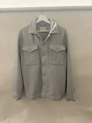 Ljusgrå overshirt med fickor (These Glory Days) - Stilren ljusgrå overshirt med klassisk krage och knäppning framtill. Två stora bröstfickor med lock och knappar ger en snygg och praktisk look. Tillverkad i ett mjukt, tjockare tyg som passar perfekt som lager. Enkel och clean design som funkar till det mesta. Storlek M men passar S också!
