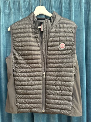 Svart dunväst från Colmar - Snygg svart dunväst från Colmar med quiltad framsida och slät baksida. Västen har hög krage, dragkedja framtill och Colmar-logga på bröstet. Perfekt att slänga över en hoodie eller tröja för extra värme. Materialet är lätt och smidigt.
