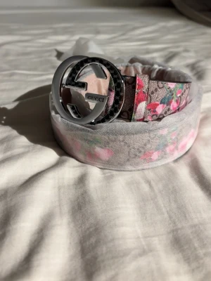Blommigt bälte  - Snyggt bälte från lager 157 med ett stort, silverfärgat GG-spänne. Bältet har ett blommigt mönster i rosa och grönt på beige botten. Perfekt accessoar för att lyfta vilken outfit som helst.