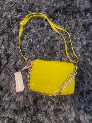 Ulrika handväska med kedja - Trendig neongul handväska i quiltad design med justerbar axelrem och en chunky guldfärgad kedja. Väskan har dragkedja baktill och en stilren look som sticker ut. Perfekt för dig som vill ha en färgklick till outfiten. Materialet är glansigt konstläder.
