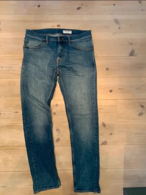 Blå jeans från Tiger of Sweden - Säljer ett par klassiska blå jeans från Tiger of Sweden. De har en smal passform, fem fickor och snygga slitningar på låren. Jeansen har gylf med knapp och dragkedja samt detaljerade sömmar. Perfekta till en avslappnad stil.