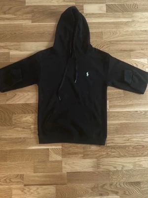 Ralph lauren hoodie - Svart hoodie med huva och dragsnören, samt korta ärmar. Liten vit logga broderad på bröstet. Perfekt för en avslappnad och cool stil. Mjuk bomullskänsla och ribbad nederkant.
