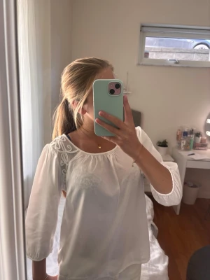 Vit blus med broderi  - Säljer en vit blus med fina broderade detaljer vid axeln och rund halsringning. Blusen har trekvartsärmar och är gjord i ett lätt, luftigt material som känns skönt mot huden. Perfekt för en stilren och fräsch look.
