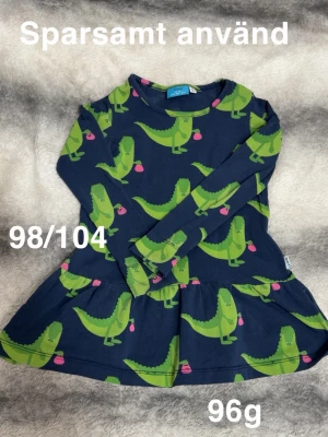 Mörkblå topp med dinosauriemönster - Mörkblå långärmad topp med gröna dinosaurier och små rosa detaljer. Tillverkad i mjuk bomull och har en rund halsringning. Perfekt för dig som gillar färgglada och lekfulla prints.