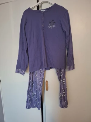 Lila pyjamas med blommönster - Mysig lila pyjamas med långärmad tröja och knappar framtill. Tröjan har ett tryck med texten 'love' och små blommor på bröstet. Byxorna är lila med vita ränder och blommönster samt resår i midjan för extra komfort.