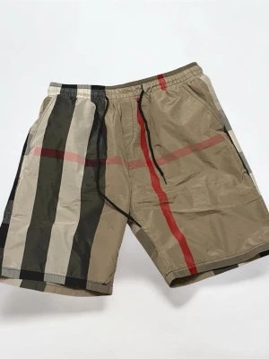 Burberry shorts size M  - Snygga beige shorts med stora rutor i svart, vitt och rött. Shortsen har elastisk midja med svart snörning och är tillverkade i ett lätt syntetmaterial. Perfekta för sommaren och ger en trendig look med sitt rutiga mönster.