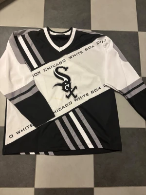 Chicago White Sox tröja CMP Team Sport - Säljer en svart och vit långärmad sporttröja från CMP Team Sport med Chicago White Sox-tryck och logga framtill. Tröjan har ränder i grått och vitt, v-ringad hals och är tillverkad i ett lätt och ventilerande syntetmaterial. Perfekt för sport eller streetwear.