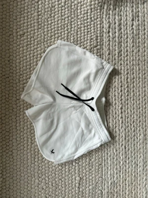 Vita sportiga shorts med snörning - Snygga vita shorts med svart snörning i midjan och en liten broderad logga på benet. De har en sportig look med rundade kanter och mjukt material som passar perfekt till sommaren eller träning.