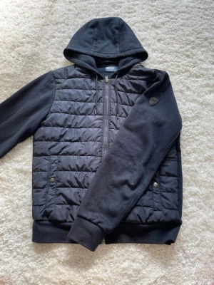 Marinblå Ralph lauren hybrid jacka - Snygg marinblå hybridjacka med huva och dragkedja framtill. Jackan har quiltad framsida, mjuka ärmar och ribbade muddar. Två fickor med tryckknappar och snörning i huvan ger extra stil och funktion. Storlek: S (passar S/M)