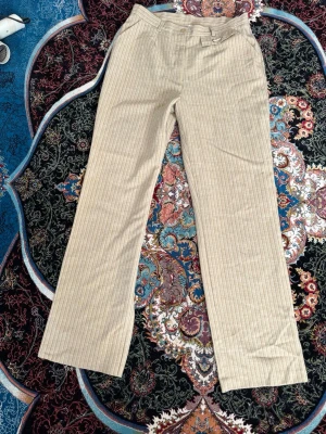 Beige randiga linnebyxor - Snygga beige linnebyxor med tunna vita ränder. Byxorna har raka ben, bälteshällor och en dekorativ ficka framtill. Tillverkade i ett luftigt linnematerial som passar perfekt till sommaren. Stängs med knapp och dragkedja.