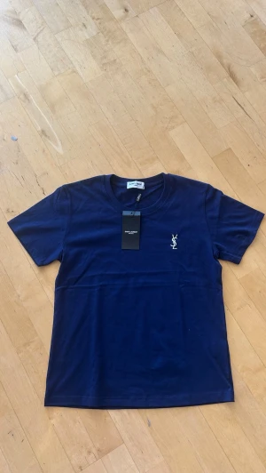Marinblå t-shirt från Saint Laurent - Snygg marinblå t-shirt från Saint Laurent med klassisk passform och korta ärmar. T-shirten har en vit YSL-logga broderad på bröstet och är gjord i mjuk bomull. Perfekt till jeans eller shorts för en clean look.