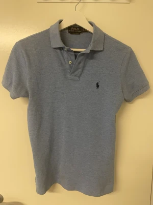 Ljusblå pikétröja från Polo Ralph Lauren - Klassisk ljusblå pikétröja från Polo Ralph Lauren med korta ärmar och krage. Tröjan har två knappar vid halsen och den ikoniska loggan broderad på bröstet. Perfekt till jeans eller shorts för en avslappnad men snygg look.