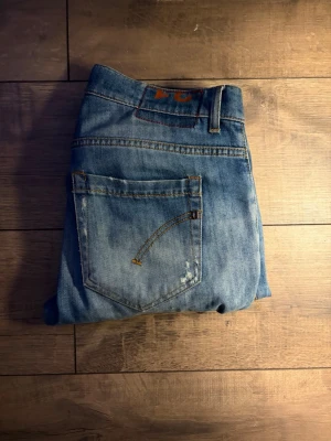 Dondup George jeans  - Säljer ett par klassiska blå jeans med raka ben och snygga slitningar framtill. Jeansen har fem fickor och bälteshällor, samt en bakficka med dekorativ söm. Perfekta för en avslappnad och trendig look.