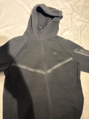 Svart Nike hoodie med dragkedja - Snygg svart hoodie från Nike med hel dragkedja framtill och huva. Har en diskret Nike-logga på bröstet och en ficka med dragkedja på ärmen. Tillverkad i mjukt material, perfekt för chill eller träning.